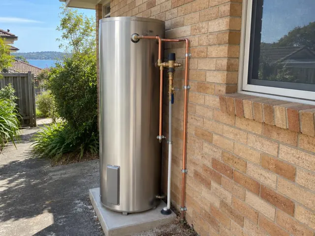 Outdoor hot water cylinder beside a brick wall and window with copper pipework in Narrabeen