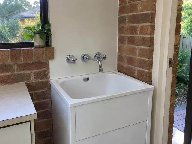 White laundry sink with wall tap set beside a brick wall and window in Mona Vale